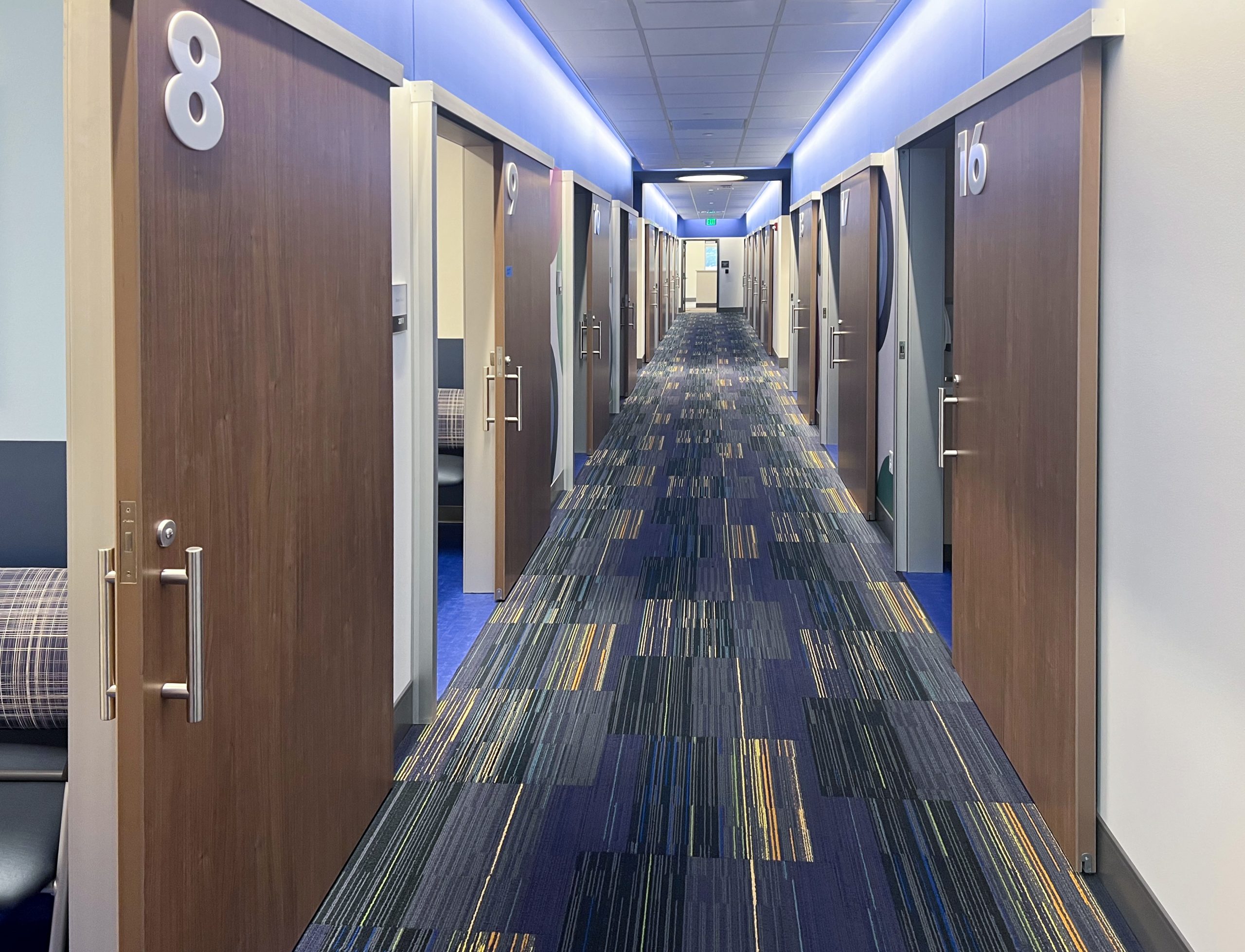 Long hallway of modern doors on both sides, numbered rooms visible (e.g., 6 and 8), blue patterned carpet and cool blue lighting