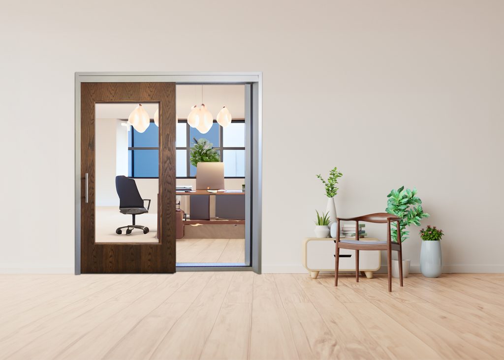 Office workspace visible through a wooden-framed glass door, with a desk, chair, hanging lights, and green plants nearby.