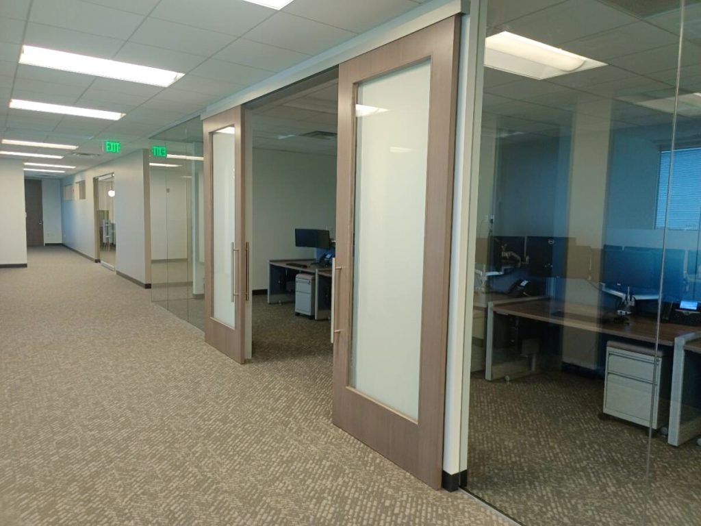 Empty office corridor with glass-walled private offices, wood-framed doors, and fluorescent lighting, exit signs visible.